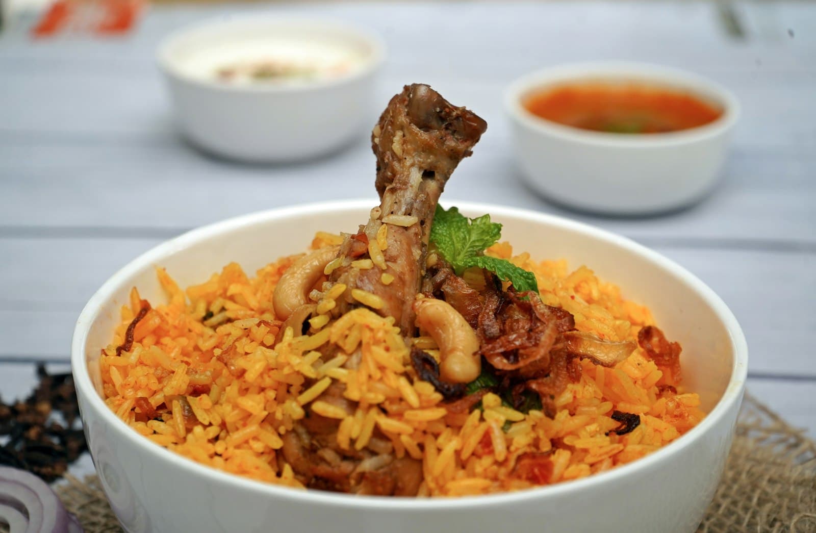 A bowl of chicken biriyani with a drumstick on top, raita and gravy behind
