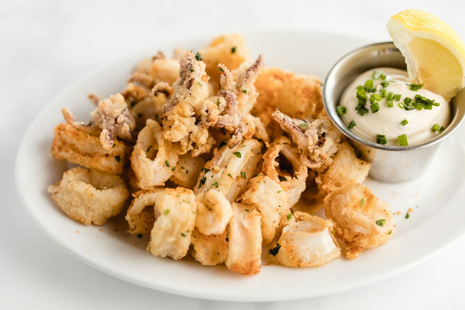Golden fried calamari on a white plate with a dipping sauce and lemon